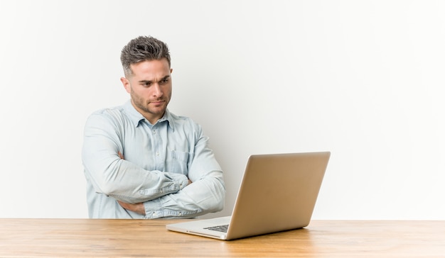 Hombre guapo joven que trabaja con su computadora portátil que se siente seguro, cruzando los brazos con determinación.