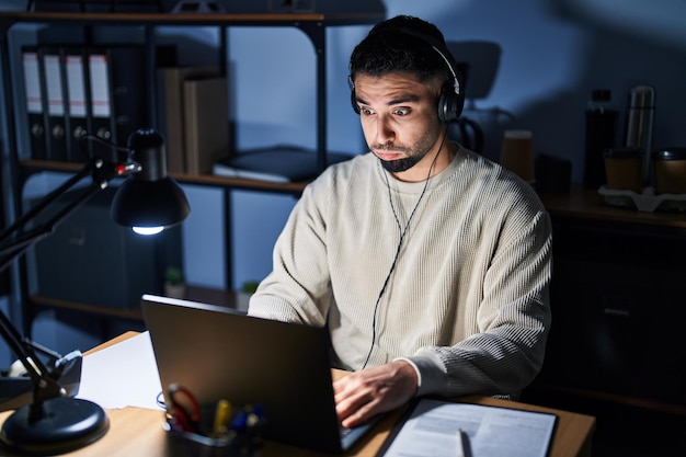 Hombre guapo joven que trabaja usando computadora portátil en la noche hinchando las mejillas con cara divertida boca inflada con expresión loca de aire