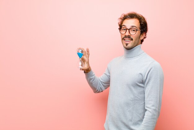 Hombre guapo joven con un reloj de arena temporizador contra pared plana rosa