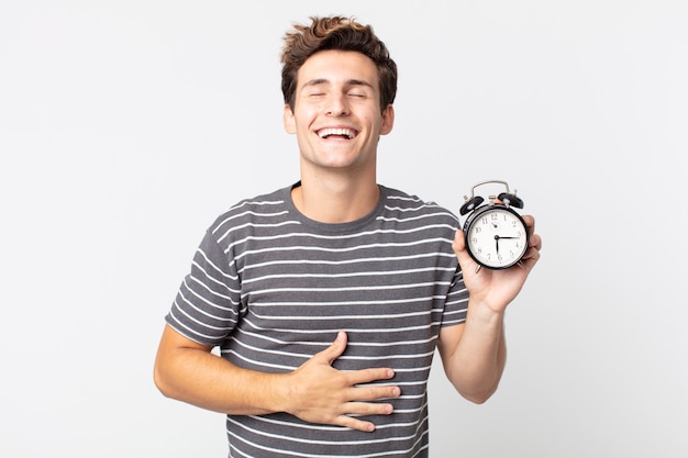 Hombre guapo joven riendo a carcajadas de una broma hilarante y sosteniendo un reloj despertador