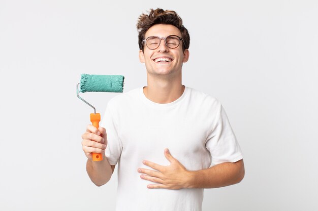 Hombre guapo joven riendo a carcajadas de una broma hilarante y sosteniendo un rodillo de pintura