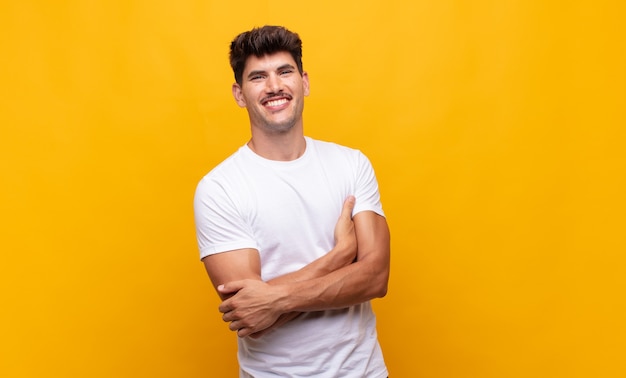 Hombre guapo joven riendo felizmente con los brazos cruzados, con una pose relajada, positiva y satisfecha