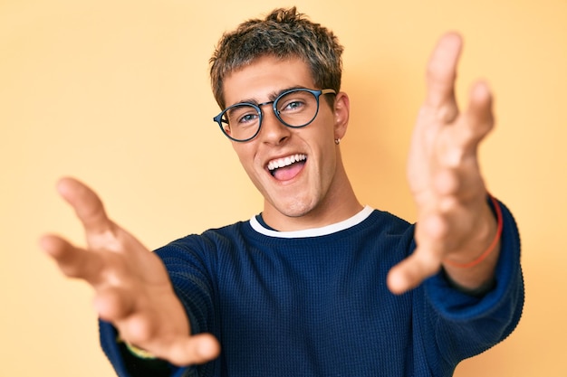 Hombre guapo joven con ropa informal y gafas mirando a la cámara sonriendo con los brazos abiertos para abrazar expresión alegre abrazando la felicidad