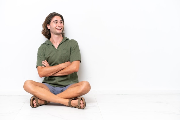 Foto hombre guapo joven sentado en el suelo aislado sobre fondo blanco con los brazos cruzados y feliz