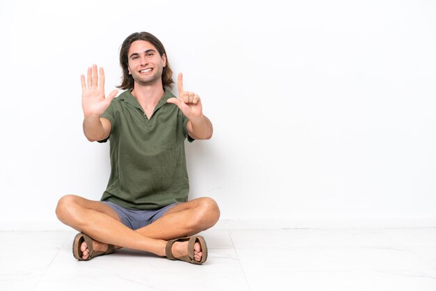 Foto hombre guapo joven sentado en el suelo aislado sobre fondo blanco contando siete con los dedos