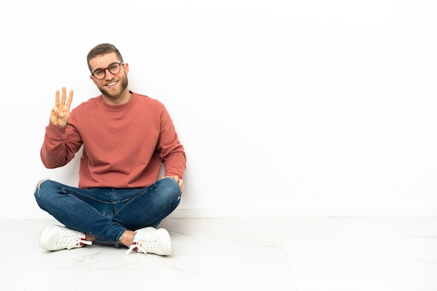 Hombre guapo joven sentado en el suelo feliz y contando tres con los dedos