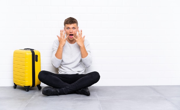 Hombre guapo joven sentado en el suelo con una maleta con expresión facial sorpresa