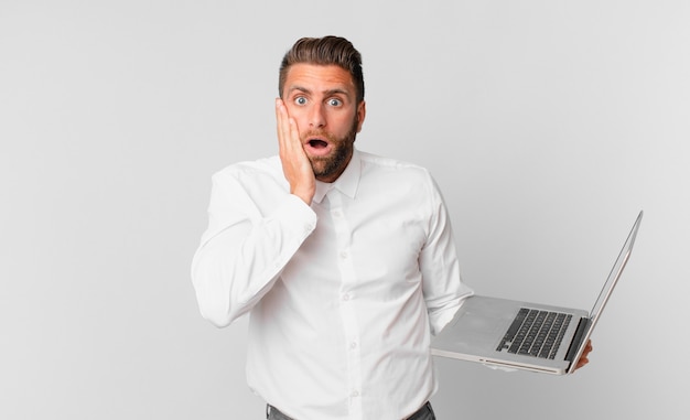 Hombre guapo joven sintiéndose conmocionado y asustado y sosteniendo una computadora portátil