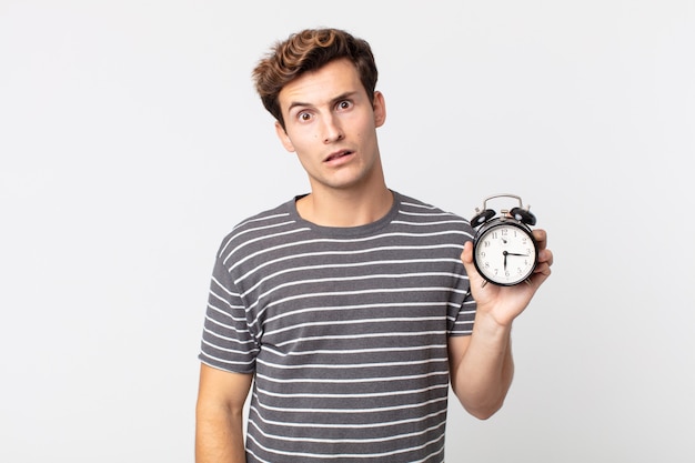 Hombre guapo joven sintiéndose desconcertado y confundido y sosteniendo un reloj despertador