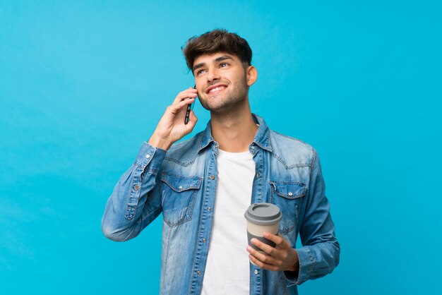 Hombre guapo joven sobre azul aislado sosteniendo café para llevar y un móvil