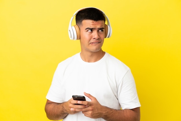 Hombre guapo joven sobre fondo amarillo aislado escuchando música con un móvil y pensando