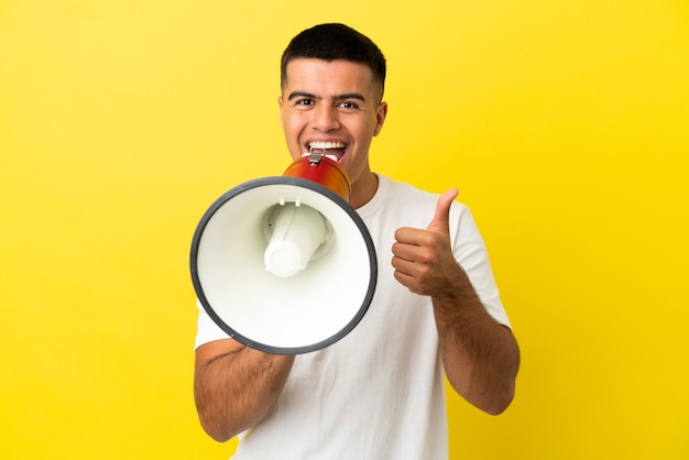 Hombre guapo joven sobre fondo amarillo aislado gritando a través de un megáfono para anunciar algo y con el pulgar hacia arriba