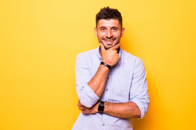 Hombre guapo joven sobre fondo amarillo aislado mirando confiado en la cámara sonriendo con los brazos cruzados y la mano levantada en la barbilla. Pensando en positivo.