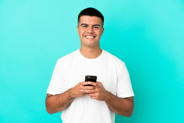 Foto hombre guapo joven sobre fondo azul aislado mirando a la cámara y sonriendo mientras usa el móvil