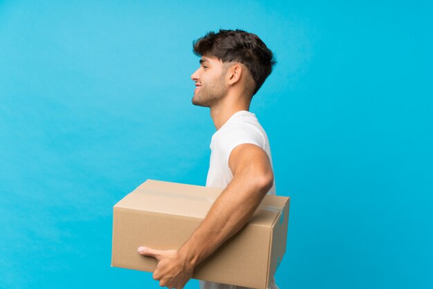 Hombre guapo joven sobre fondo azul aislado sosteniendo una caja para moverlo a otro sitio en posición lateral