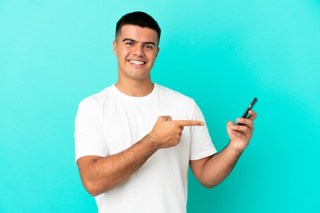 Hombre guapo joven sobre fondo azul aislado mediante teléfono móvil y apuntando