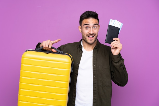 Hombre guapo joven sobre pared púrpura aislada en vacaciones con maleta y pasaporte y sorprendido