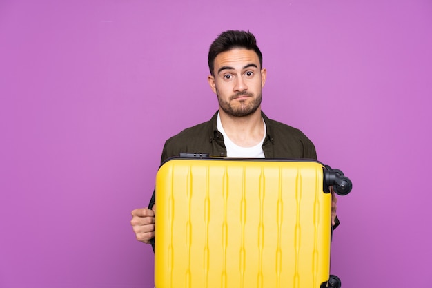 Hombre guapo joven sobre pared púrpura aislada en vacaciones con maleta de viaje