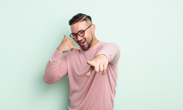 Hombre guapo joven sonriendo alegremente y apuntando a la cámara mientras hace una llamada, gesto más tarde, hablando por teléfono