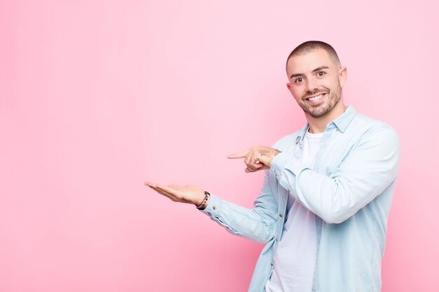 Hombre guapo joven sonriendo alegremente y apuntando para copiar espacio en la palma en el lateral, mostrando o anunciando un objeto contra la pared plana