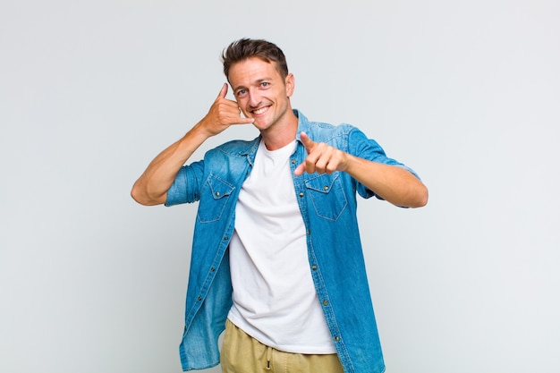 Hombre guapo joven sonriendo alegremente y apuntando hacia el frente mientras hace una llamada, gesto más tarde, hablando por teléfono