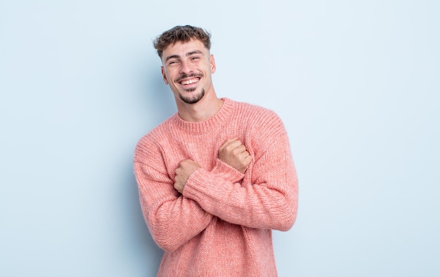 Hombre guapo joven sonriendo alegremente y celebrando, con los puños apretados y los brazos cruzados, sintiéndose feliz y positivo