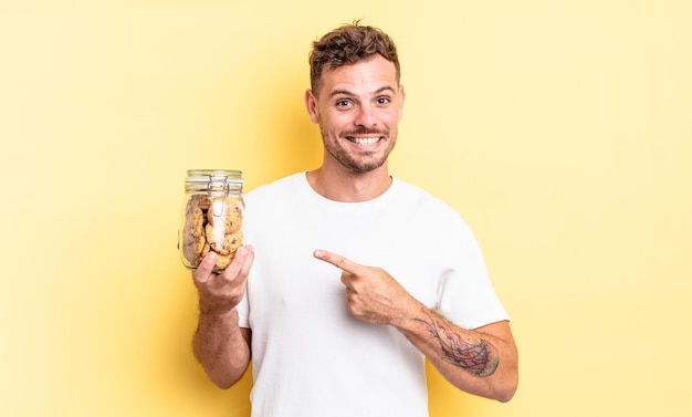 Hombre guapo joven sonriendo alegremente, sintiéndose feliz y apuntando al concepto de botella de galletas de lado