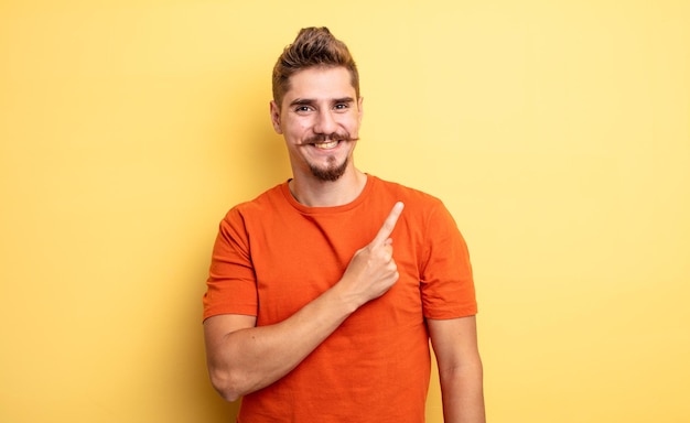 Hombre guapo joven sonriendo alegremente, sintiéndose feliz y apuntando hacia un lado y hacia arriba, mostrando objetos en el espacio de la copia. extraño concepto de bigote