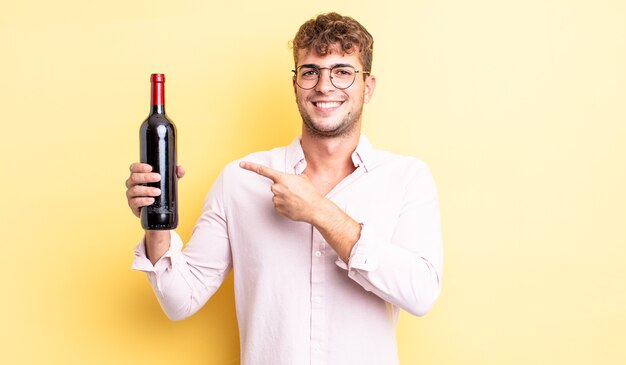 Hombre guapo joven sonriendo alegremente, sintiéndose feliz y apuntando hacia un lado. concepto de botella de vino