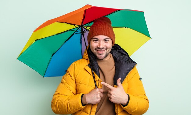 Hombre guapo joven sonriendo alegremente, sintiéndose feliz y apuntando hacia un lado. concepto de lluvia y paraguas