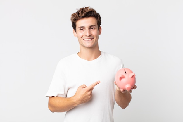 Hombre guapo joven sonriendo alegremente, sintiéndose feliz y apuntando hacia un lado y sosteniendo una alcancía