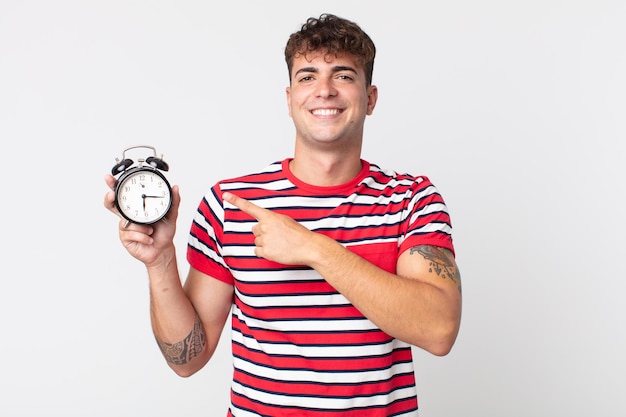 Hombre guapo joven sonriendo alegremente, sintiéndose feliz y apuntando hacia un lado y sosteniendo un despertador