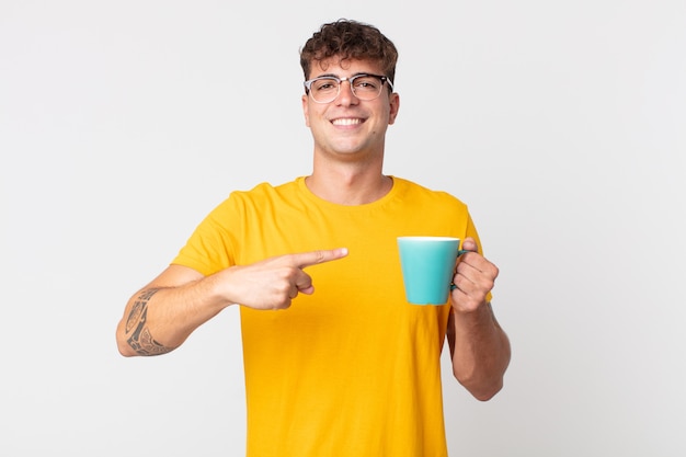 Hombre guapo joven sonriendo alegremente, sintiéndose feliz y apuntando hacia un lado y sosteniendo una taza de café