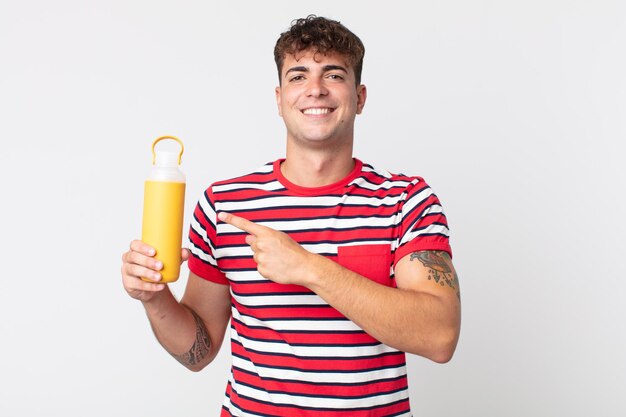 Hombre guapo joven sonriendo alegremente sintiéndose feliz y apuntando hacia un lado y sosteniendo un termo de café