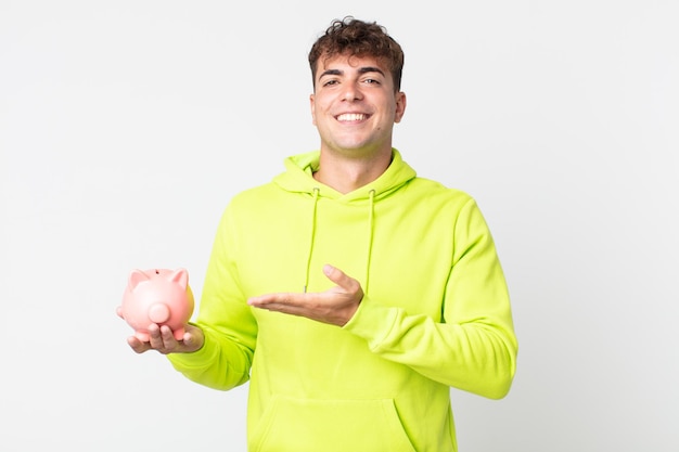 Hombre guapo joven sonriendo alegremente, sintiéndose feliz y mostrando un concepto y sosteniendo una alcancía