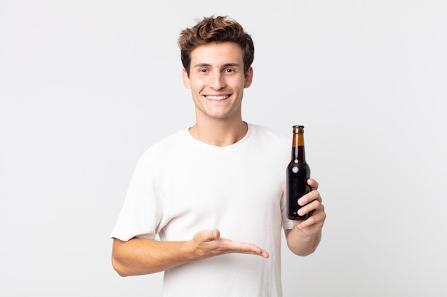 Hombre guapo joven sonriendo alegremente, sintiéndose feliz y mostrando un concepto y sosteniendo una botella de cerveza