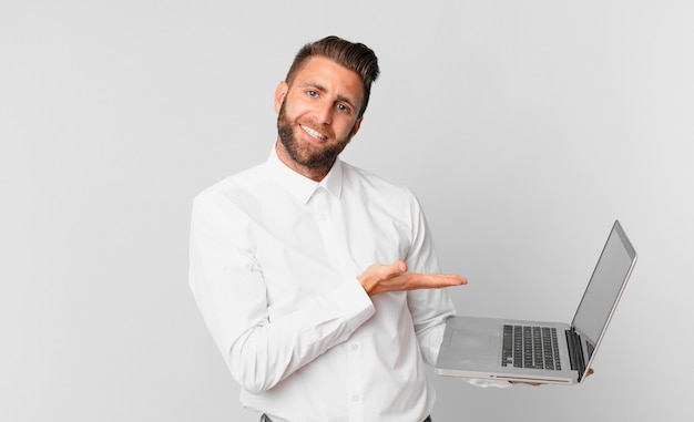 Hombre guapo joven sonriendo alegremente, sintiéndose feliz y mostrando un concepto y sosteniendo una computadora portátil