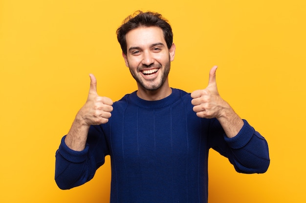 Hombre guapo joven sonriendo ampliamente mirando feliz, positivo, seguro y exitoso, con ambos pulgares hacia arriba