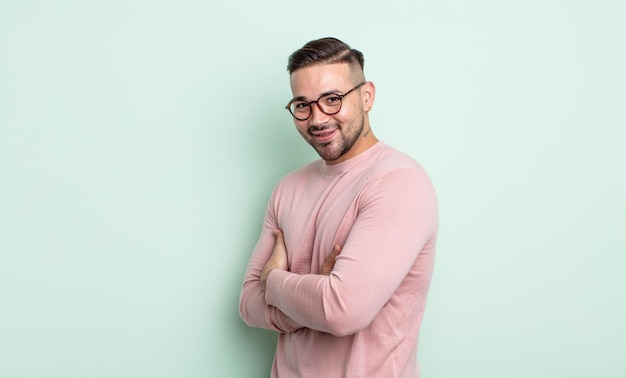 Hombre guapo joven sonriendo a la cámara con los brazos cruzados y una expresión feliz, segura y satisfecha, vista lateral