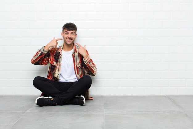 Hombre guapo joven sonriendo con confianza apuntando a su propia sonrisa amplia, actitud positiva, relajada y satisfecha sentado en el piso de cemento