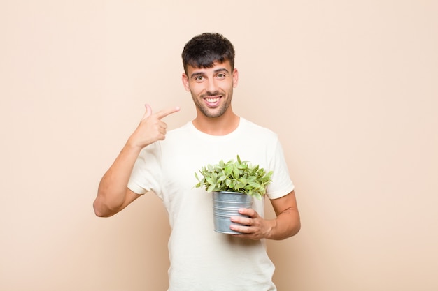 Hombre guapo joven sonriendo con confianza apuntando a su propia sonrisa amplia, actitud positiva, relajada y satisfecha sosteniendo una planta