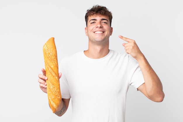 Hombre guapo joven sonriendo con confianza apuntando a su propia sonrisa amplia y sosteniendo una baguette de pan