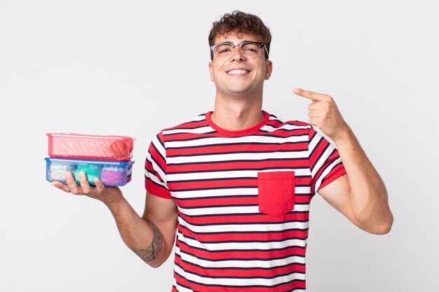 Hombre guapo joven sonriendo con confianza apuntando a su propia sonrisa amplia y sosteniendo una lonchera