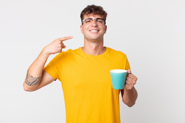 Hombre guapo joven sonriendo con confianza apuntando a su propia sonrisa amplia y sosteniendo una taza de café