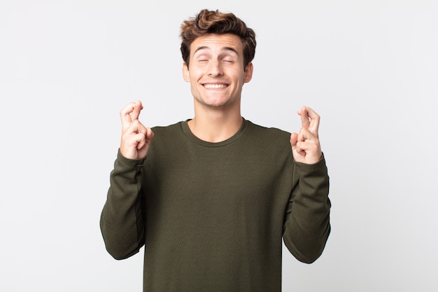 Hombre guapo joven sonriendo y cruzando ambos dedos ansiosamente, sintiéndose preocupado y deseando o esperando buena suerte