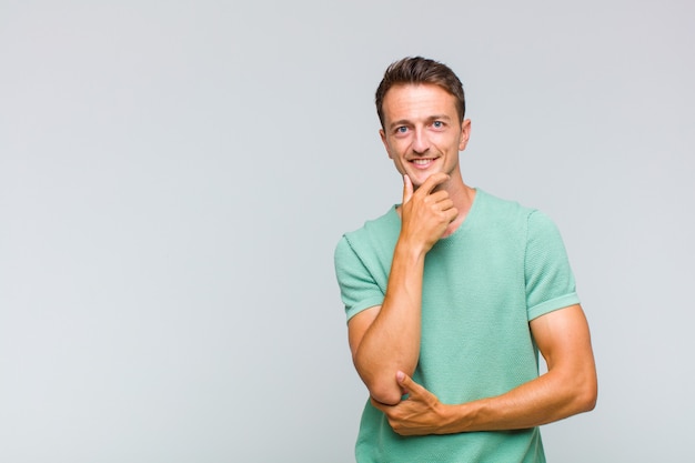 Hombre guapo joven sonriendo, disfrutando de la vida, sintiéndose feliz, amigable, satisfecho y despreocupado con la mano en la barbilla