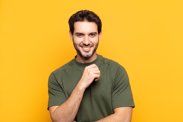 Hombre guapo joven sonriendo, disfrutando de la vida, sintiéndose feliz, amigable, satisfecho y despreocupado con la mano en la barbilla