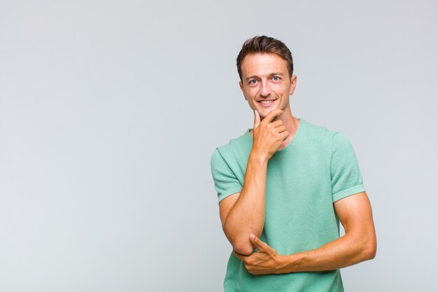 Hombre guapo joven sonriendo, disfrutando de la vida, sintiéndose feliz, amistoso, satisfecho y despreocupado con la mano en la barbilla