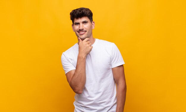 Hombre guapo joven sonriendo, disfrutando de la vida, sintiéndose feliz, amistoso, satisfecho y despreocupado con la mano en la barbilla