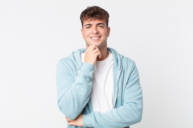 Hombre guapo joven sonriendo, disfrutando de la vida, sintiéndose feliz, amistoso, satisfecho y despreocupado con la mano en la barbilla